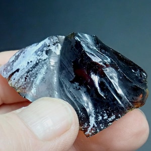 May include: A dark, iridescent obsidian rock with a rough, textured surface. The rock has a silvery sheen and a hint of red.