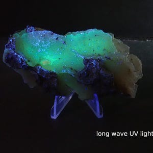 May include: A mineral specimen illuminated by long wave UV light, exhibiting bright green and blue fluorescence. The rock is set against a dark backdrop. The text "long wave UV light" is visible. The mineral is displayed on a clear plastic stand.