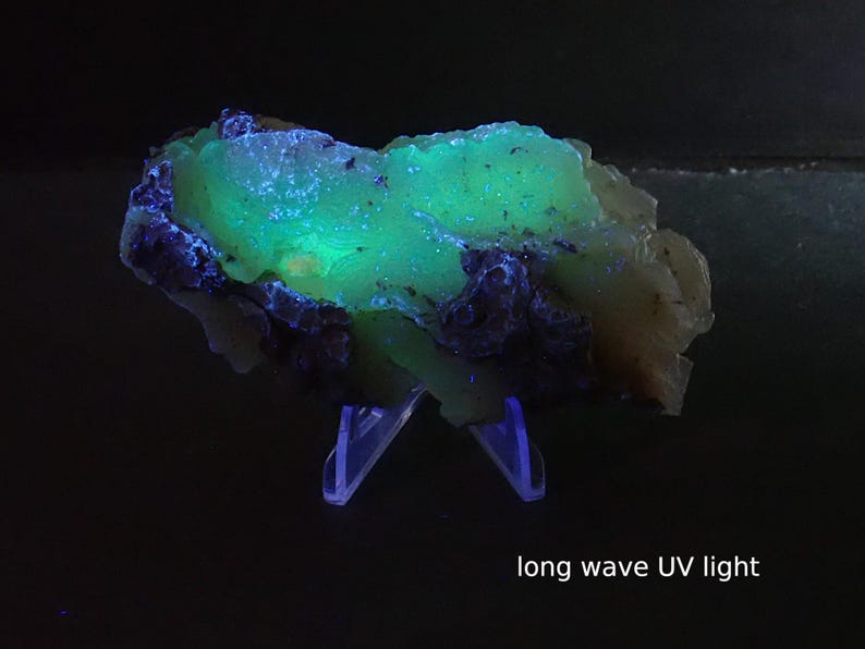 May include: A mineral specimen illuminated by long wave UV light, exhibiting bright green and blue fluorescence. The rock is set against a dark backdrop. The text "long wave UV light" is visible. The mineral is displayed on a clear plastic stand.