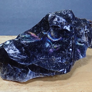 May include: A raw, dark obsidian stone with a glossy, reflective surface. The mineral features irregular, jagged edges and deep black coloration, with subtle iridescent flashes of purple, blue, and green visible across its fractured, glassy texture.