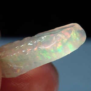 May include: A translucent white opal gemstone with iridescent green, blue, and orange flashes. The opal is rough and has a natural, irregular shape.