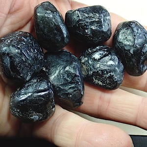 May include: A collection of seven polished, dark blue-black tumbled stones. The stones have a smooth, rounded shape with a slightly textured surface. They are held in an open hand, showcasing their natural beauty and potential for use in jewelry or crafts.