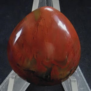 May include: A smooth, polished red jasper cabochon with brown and black swirling patterns. The stone is oval shaped and has a glossy finish.