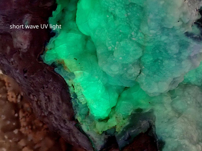 May include: A close-up of a green mineral under short wave UV light. The mineral has a bubbly texture and is glowing brightly.