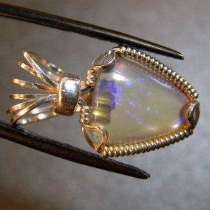 May include: A delicate gold pendant featuring a white opal gemstone with iridescent blue and purple hues. The opal is set in a twisted wire frame with a decorative bail.