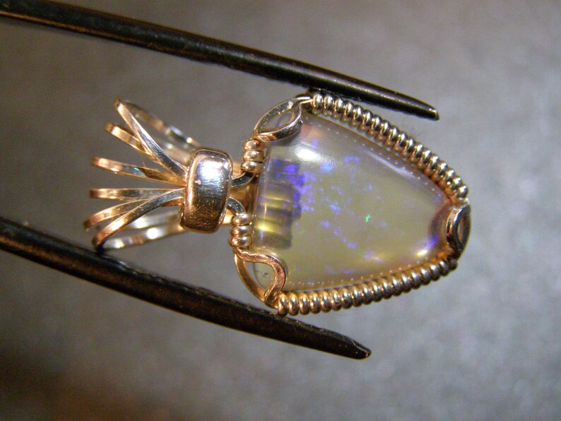 May include: A delicate gold pendant featuring a white opal gemstone with iridescent blue and purple hues. The opal is set in a twisted wire frame with a decorative bail.