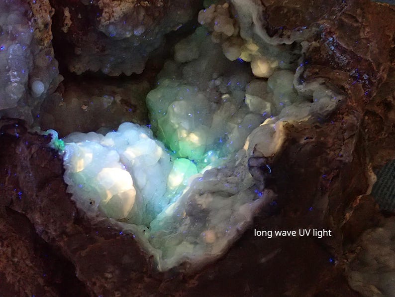 May include: A close-up of a geode with a white and green interior. The geode is illuminated with long wave UV light, causing the interior to glow a bright blue and green.