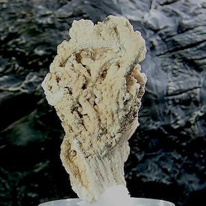 Puede incluir: Un esp&eacute;cimen mineral de color beige claro con una textura rugosa y porosa. El mineral tiene una forma vertical y alargada con una estructura cristalina. El fondo es gris oscuro moteado, proporcionando contraste con el mineral de color claro.