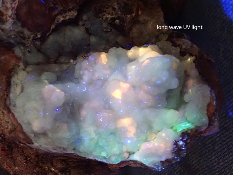 May include: A close-up of a light blue and green mineral under long wave UV light. The mineral glows brightly in shades of blue, green, and white.