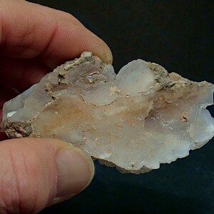 May include: A piece of raw quartz crystal held in a hand. The crystal is a mix of translucent white and tan colours, with a rough, uneven texture. The edges are jagged, and the overall shape is irregular.