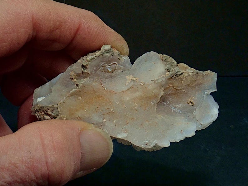 May include: A piece of raw quartz crystal held in a hand. The crystal is a mix of translucent white and tan colours, with a rough, uneven texture. The edges are jagged, and the overall shape is irregular.