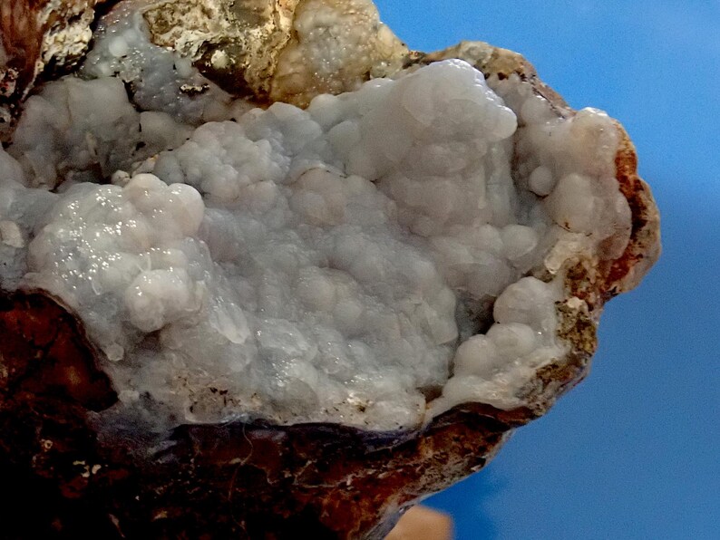 May include: A close-up of a geode with a light purple and white crystal formation. The geode is partially broken open, revealing the crystals inside. The geode is surrounded by a brown, rocky exterior.