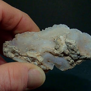 May include: A raw mineral specimen with a crystalline structure, held in a hand. The stone features a mix of white and light grey hues, with a rough, textured surface. The mineral's natural form is visible.