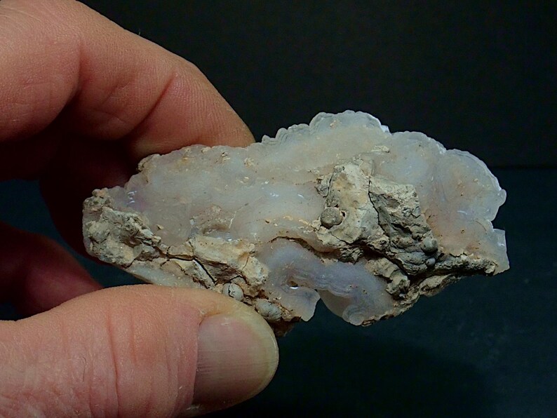 May include: A raw mineral specimen with a crystalline structure, held in a hand. The stone features a mix of white and light grey hues, with a rough, textured surface. The mineral's natural form is visible.