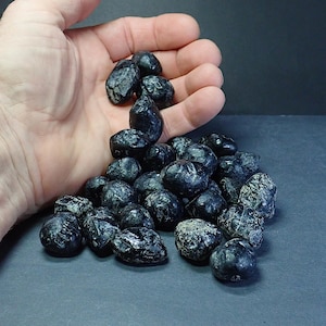 May include: A collection of small, polished black stones with irregular shapes and subtle white veining. The stones have a smooth, reflective surface texture and are grouped together in a pile, displaying various dark gray and black color tones.
