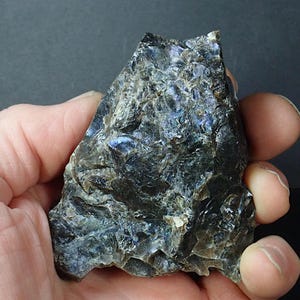 May include: A hand holds a rough, dark-colored mineral specimen. The stone has a layered, textured appearance with iridescent blue and purple flashes. The overall color is a mix of dark gray and black.