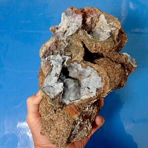 May include: A large, irregularly shaped rock with a rough, brown exterior. The rock has a cavity that reveals a cluster of white and light blue crystals.