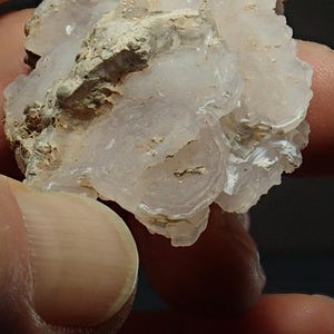 May include: A close-up of a light pink and white mineral specimen. The mineral has a layered, circular pattern with a rough, textured surface. The specimen is held in a person's hand.