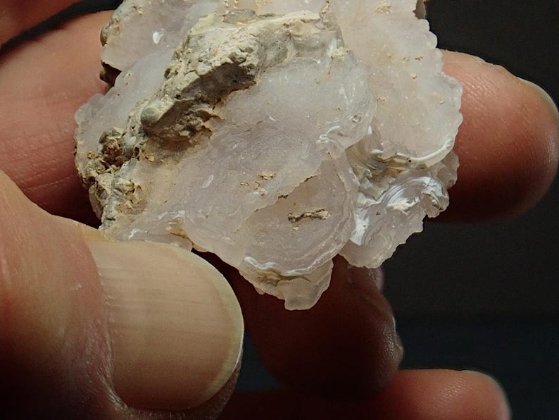 May include: A close-up of a light pink and white mineral specimen. The mineral has a layered, circular pattern with a rough, textured surface. The specimen is held in a person's hand.