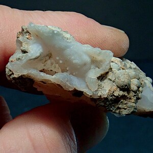 May include: A close-up of a natural mineral specimen. The stone features a light blue, crystalline interior within a rough, beige exterior. The specimen is held between fingers, showcasing its texture and colour variations. The overall shape is irregular and organic.