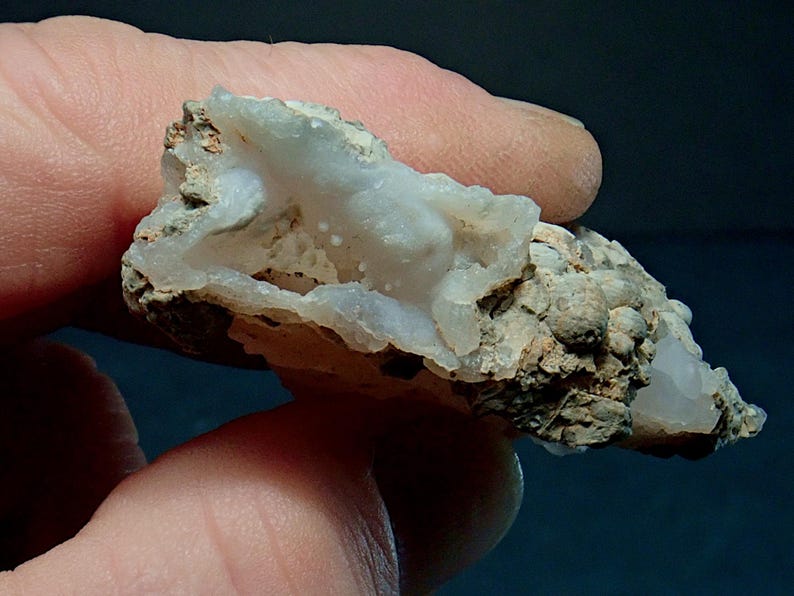 May include: A close-up of a natural mineral specimen. The stone features a light blue, crystalline interior within a rough, beige exterior. The specimen is held between fingers, showcasing its texture and colour variations. The overall shape is irregular and organic.