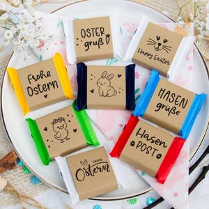 Könnte beinhalten: Draufsicht auf Oster-Schokoriegel auf einem weißen Teller. Jeder Riegel ist in braunes Papier gewickelt und mit verschiedenen Designs versehen, darunter ein Hase, ein Küken und die Worte "Happy Easter" und "Frohe Ostern". Die Riegel sind von Dekorationen umgeben.