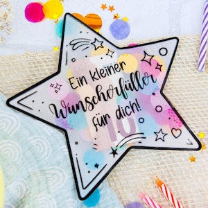 May include: A star-shaped decoration with the text "Ein kleiner Wunschfüller für dich!" in black script, surrounded by colorful confetti, candles, and ribbons. The star has a black outline and a watercolor-like design.