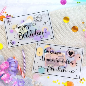 Op de afbeelding: Twee verjaardagskaarten met de woorden "happy Birthday" en "Ein kleiner Wunscherfüller für dich" in zwart schrift. De kaarten zijn versierd met kleurrijke confetti, kaarsen en linten. Een feestelijke scène.