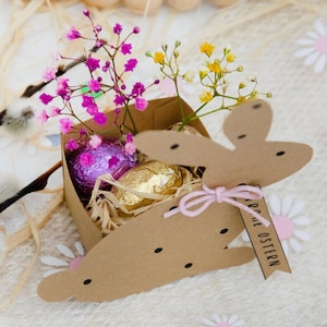 May include: A decorative Easter-themed arrangement featuring a brown cardboard carrot-shaped box with a pink bow and a tag that reads "Frohe Ostern". The box contains foil-wrapped chocolate eggs and is decorated with pink and yellow flowers and sprigs.
