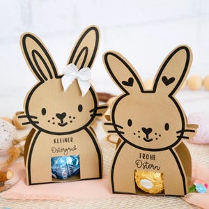 May include: Two brown bunny-shaped gift boxes with black outlines and details. One has a white bow, the other has heart-shaped accents. Each holds a wrapped chocolate. Text on the boxes reads "Kleiner Ostergruß" and "Frohe Ostern".