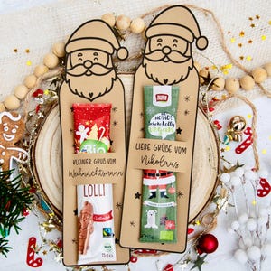 Könnte beinhalten: Zwei beigefarbene Papphalter in Weihnachtsmannform, die jeweils einen Schokoladenlutscher enthalten. Der linke trägt die Aufschrift Kleiner Gruß vom Weihnachtsmann, der rechte Liebe Grüße vom Nikolaus, beide mit weihnachtlichen Motiven.