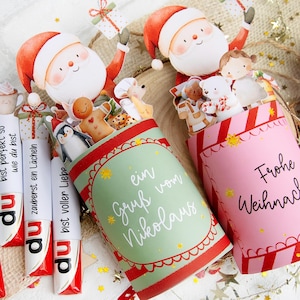 Może przedstawiać: Świąteczne pudełka na prezenty i opakowania po cukierkach z Mikołajem i świątecznymi postaciami. Pudełka są zielone i różowe, z niemieckim tekstem. Opakowania po cukierkach mają niemiecki tekst.