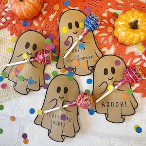 May include: Four tan paper ghost cutouts, each holding a round lollipop. The ghosts feature black printed faces and text including Happy Halloween, BOOOH!, and SÜBES ER SAURES. Two small orange pumpkins are placed among the ghost cutouts.