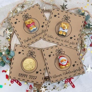 Può includere: Quattro biglietti di Natale quadrati con disegni festosi. Ogni biglietto presenta un diverso ornamento di cioccolato: un orsacchiotto, una moneta e un Babbo Natale. I biglietti sono decorati con stelle e il testo "Happy 2027".