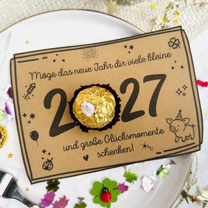 Op de afbeelding: Een rechthoekige bruine kaart met zwarte tekst 2027 en Duitse zinnen, met in het midden een in goudfolie verpakte chocolade. De kaart bevat illustraties van een varken, vuurwerk en sterren, gepresenteerd op een wit bord.