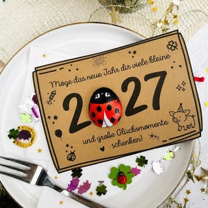 May include: A rectangular tan card featuring the text "M&ouml;ge das neue Jahr dir viele kleine 2027 und gro&szlig;e Gl&uuml;cksmomente schenken!" with a painted ladybug replacing the zero, alongside illustrations of a pig, fireworks, and stars.