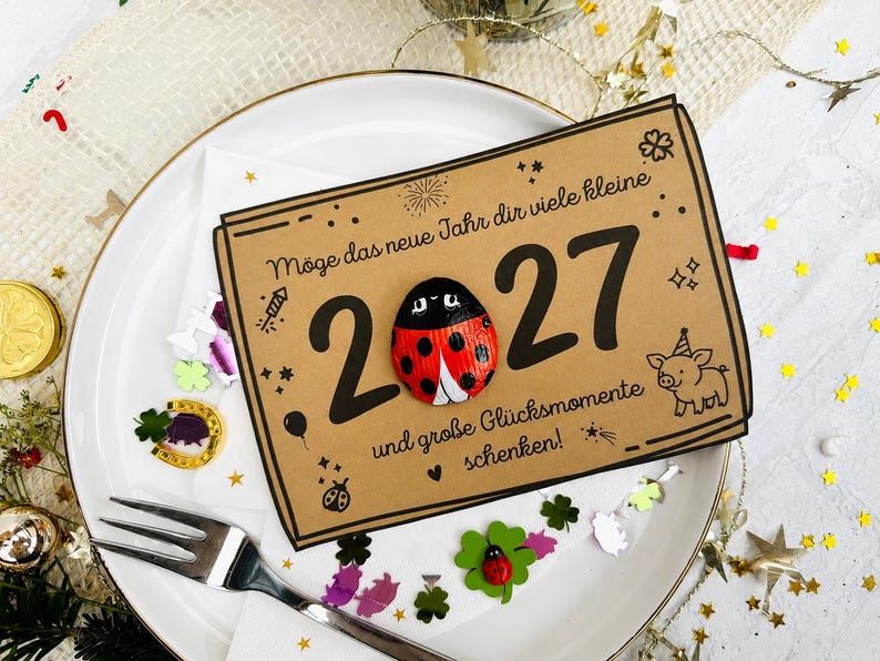 May include: A rectangular tan card featuring the text "M&ouml;ge das neue Jahr dir viele kleine 2027 und gro&szlig;e Gl&uuml;cksmomente schenken!" with a painted ladybug replacing the zero, alongside illustrations of a pig, fireworks, and stars.