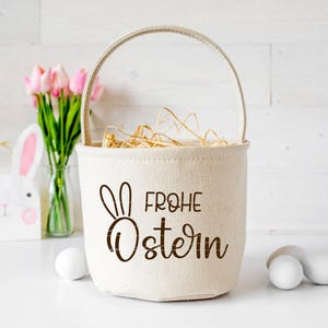 May include: A white canvas Easter basket with a brown bunny and the text "Frohe Ostern" in brown. The basket is filled with straw and is sitting on a white surface. There are pink tulips in a glass vase and a white bunny figurine in the background.