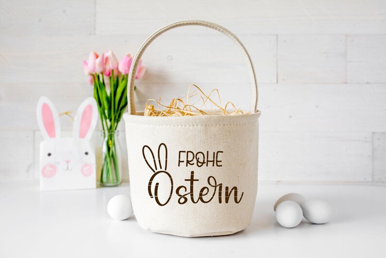 May include: A white canvas Easter basket with a brown bunny and the text "Frohe Ostern" in brown. The basket is filled with straw and is sitting on a white surface. There are pink tulips in a glass vase and a white bunny figurine in the background.