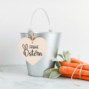 May include: A silver metal bucket with a wooden heart-shaped tag that says "Frohe Ostern" in black lettering. The tag has a small bunny rabbit illustration. The bucket is filled with orange carrots and green leafy stems.