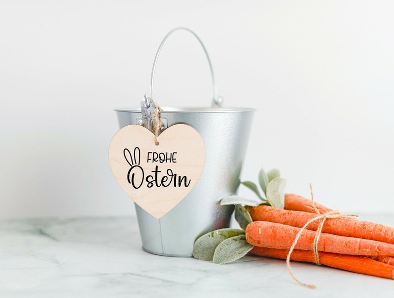 May include: A silver metal bucket with a wooden heart-shaped tag that says "Frohe Ostern" in black lettering. The tag has a small bunny rabbit illustration. The bucket is filled with orange carrots and green leafy stems.
