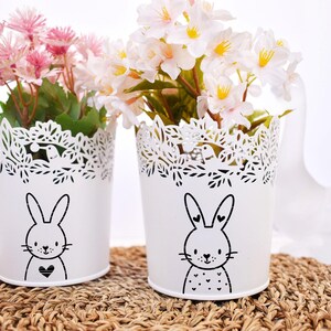 May include: Two white metal flower pots with a bunny design. The pots have a decorative, cut-out rim and are filled with pink and white flowers. They sit on a woven mat.