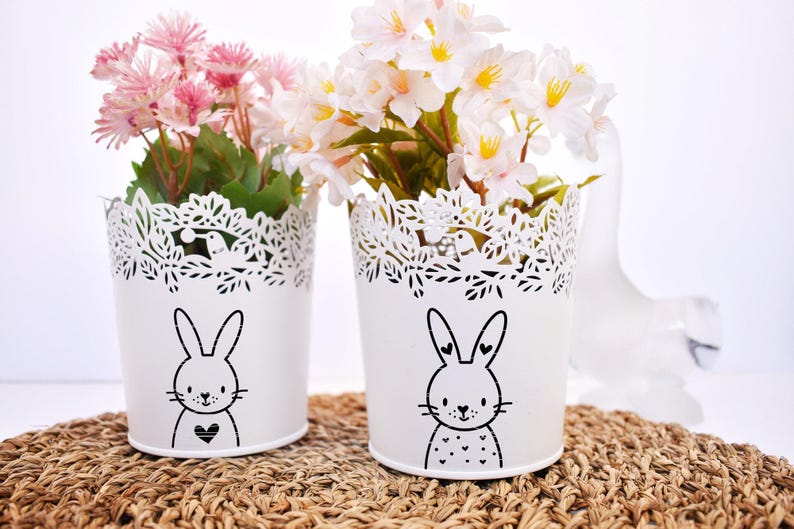 May include: Two white metal flower pots with a bunny design. The pots have a decorative, cut-out rim and are filled with pink and white flowers. They sit on a woven mat.