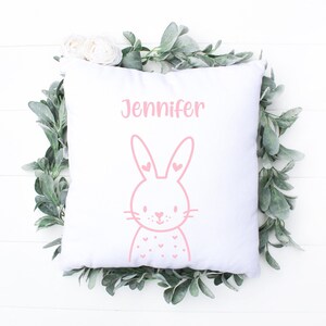 May include: White square pillow with a pink bunny illustration and the name "Jennifer" printed above it. The bunny has heart-shaped details. The pillow is surrounded by a wreath of green leaves and white flowers.