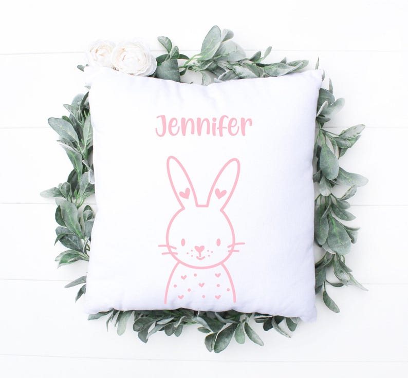 May include: White square pillow with a pink bunny illustration and the name "Jennifer" printed above it. The bunny has heart-shaped details. The pillow is surrounded by a wreath of green leaves and white flowers.