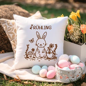 May include: White pillow with a brown bunny rabbit and the word "FR&Uuml;HLING" printed on it. The bunny is holding an egg and there are flowers and other eggs around it. The pillow is sitting on a white surface with a basket of colorful eggs in the foreground.