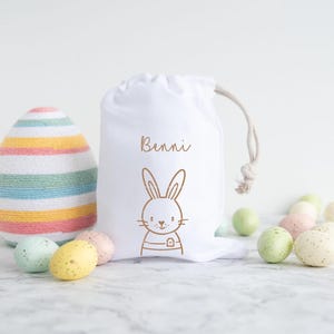 May include: White fabric Easter gift bag with a bunny and the name "Bennie" printed on it. The bag is filled with pastel colored Easter eggs.