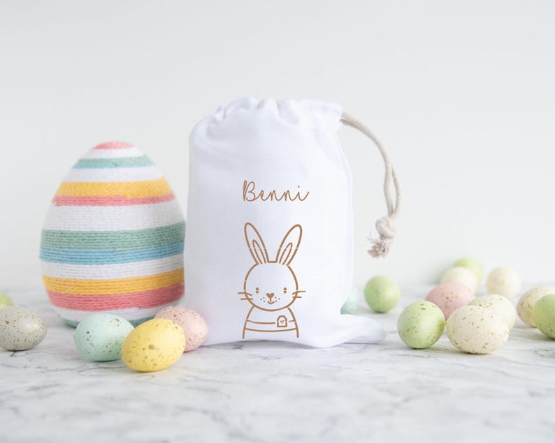 May include: White fabric Easter gift bag with a bunny and the name "Bennie" printed on it. The bag is filled with pastel colored Easter eggs.
