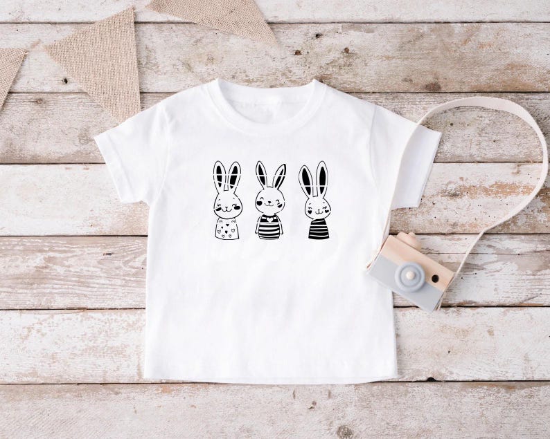 Op de afbeelding: Wit T-shirt met een zwart-witte afbeelding van drie konijnen die verschillende outfits dragen. De konijnen dragen gestreepte shirts, een stippenoverhemd en een effen shirt.