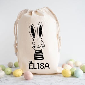May include: A white canvas drawstring bag with a black and white bunny illustration and the name "ELISA" printed on the front. The bag is filled with pastel coloured Easter eggs.